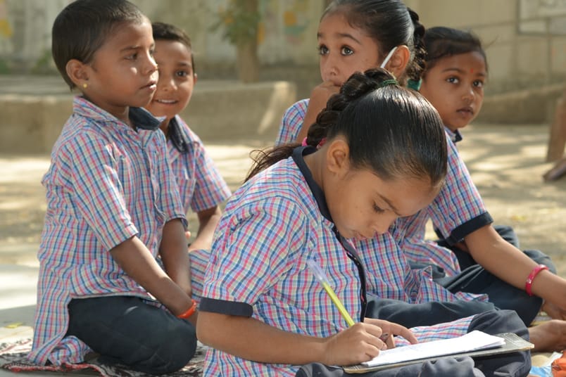 A group of children in school uniform sitting on the ground, writing in notebooks