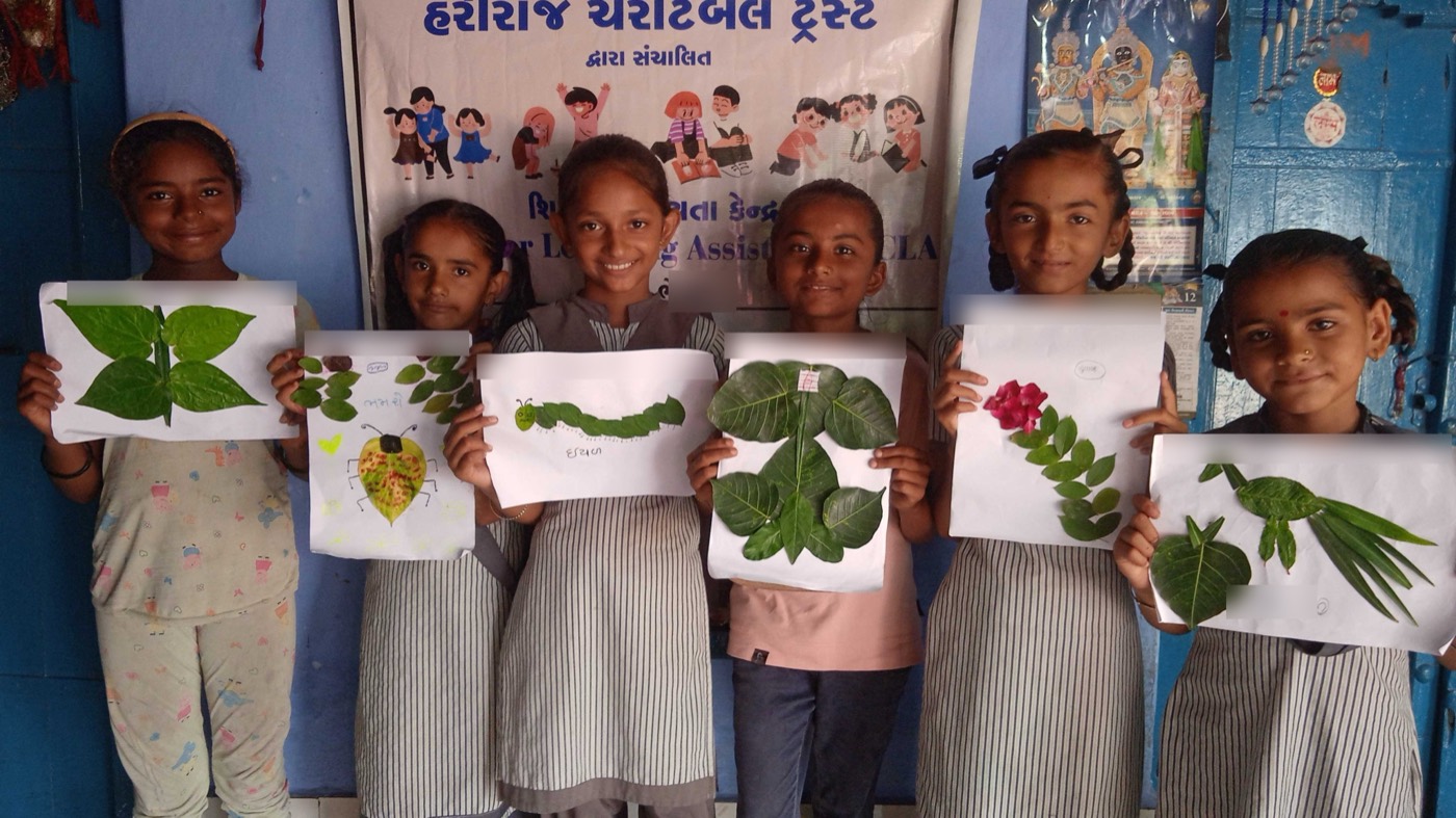 Six smiling girls standing in a row at a learning centre, each holding up a picture made from leaves — a butterfly, a caterpillar, a bird