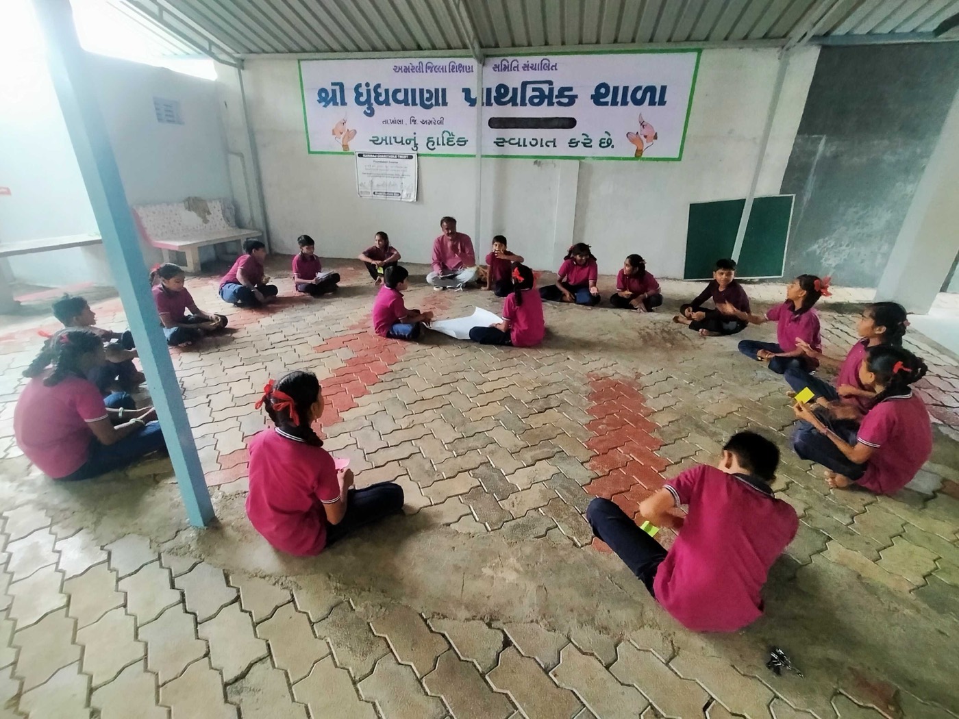 Schoolchildren in uniform sitting in a wide circle on the floor of a primary school's covered yard, with a facilitator among them
