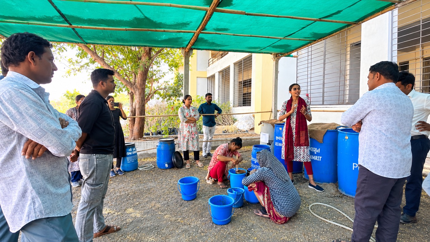 A group of farmers and field staff standing under a shade net around blue drums and buckets while a trainer explains how a bio-input is prepared