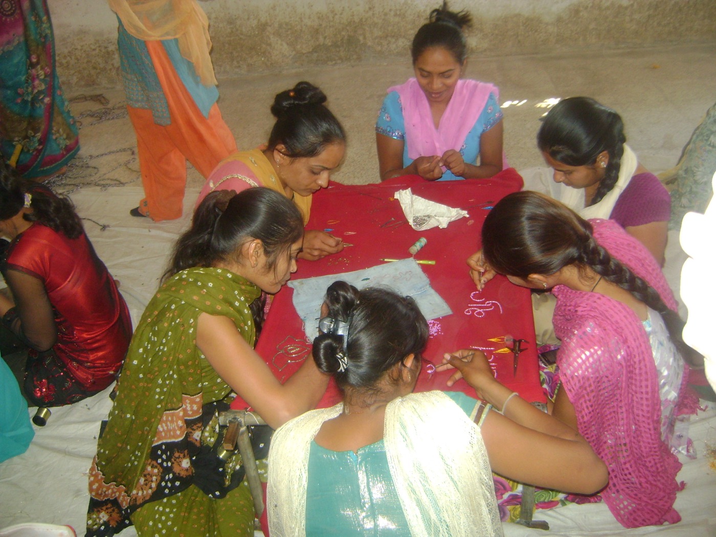 Young women seated around a red cloth spread on the floor, each working embroidery into it with a needle