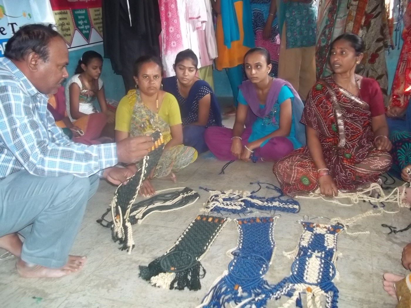 A trainer kneeling beside finished macramé pieces laid out on the floor, with women seated in a row watching