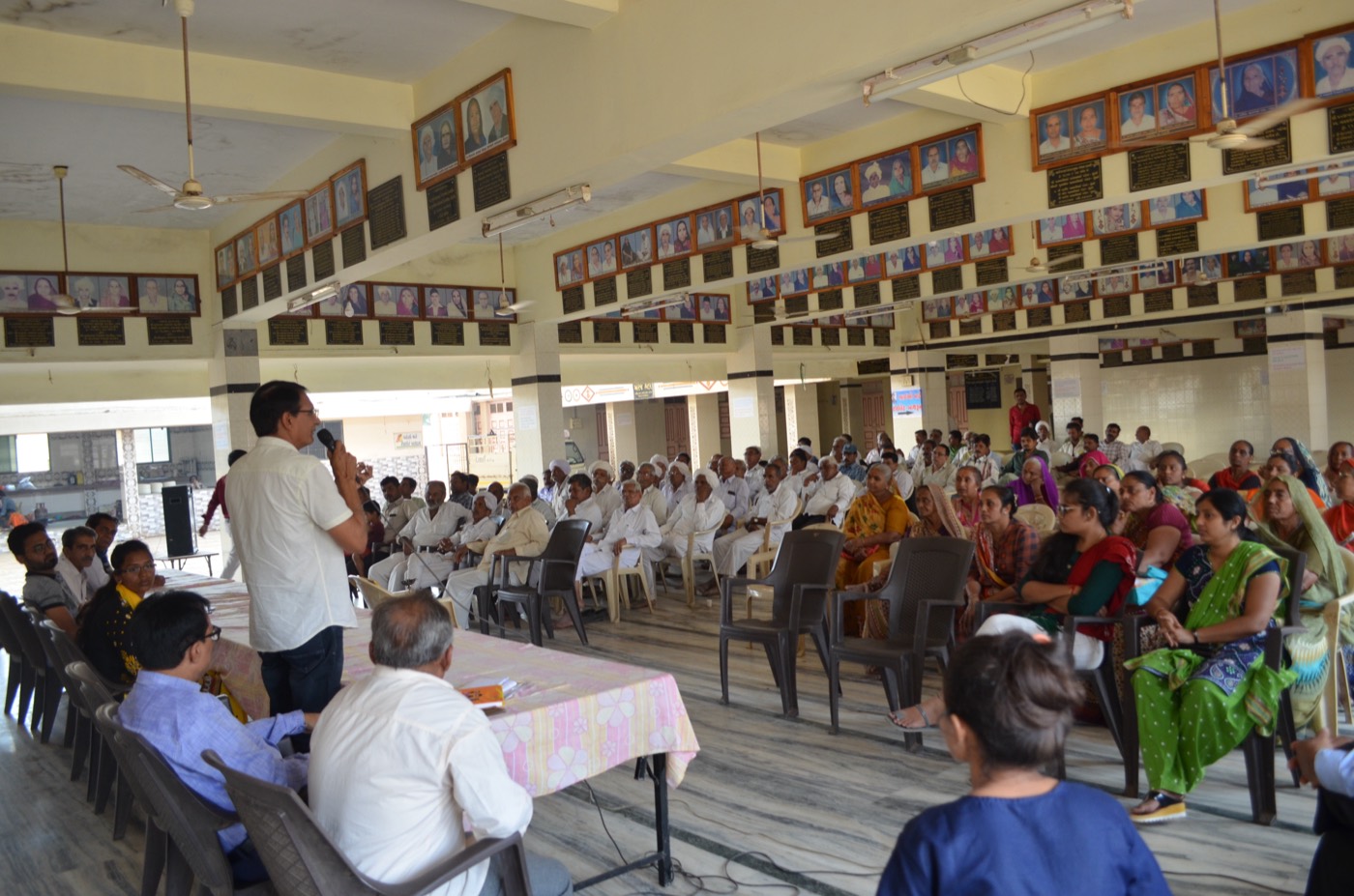 A trainer with a microphone addressing a hall of seated farmers, men on one side and women on the other