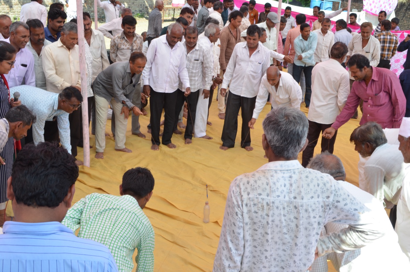 A large group of farmers standing in a ring on a mat, each holding a string attached to one bottle, working together to move it