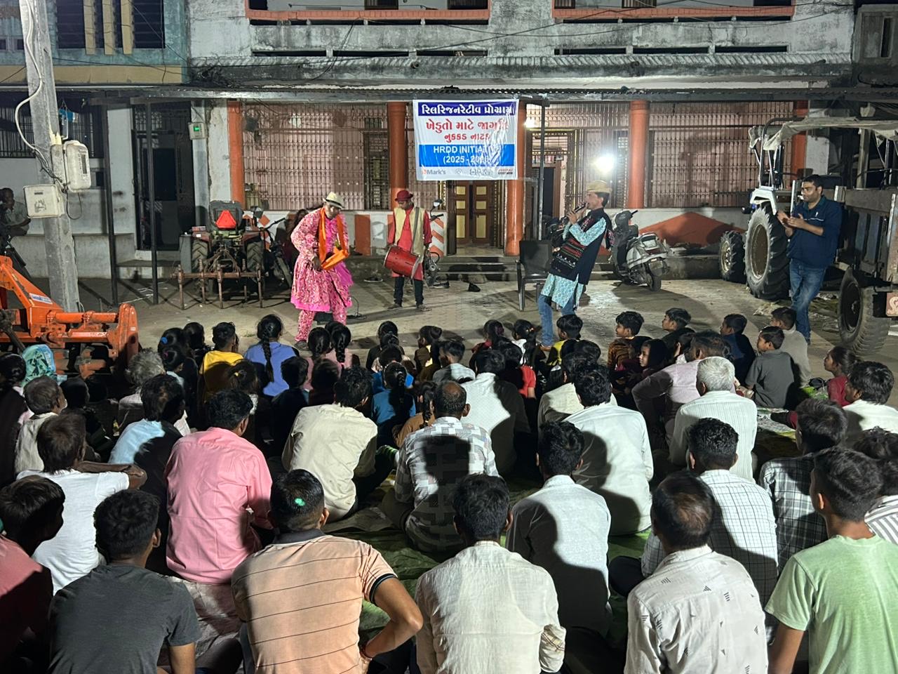 Villagers seated on the ground at night watching three performers in bright costumes, with a drummer, in front of a house