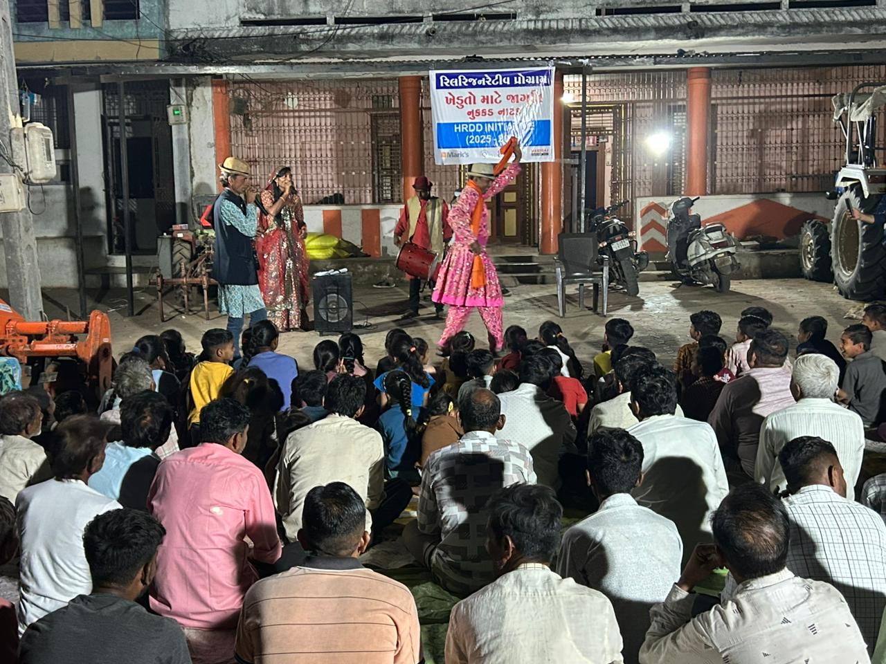 Performers in costume acting out a scene before a seated village audience, under a banner announcing an awareness street play for farmers